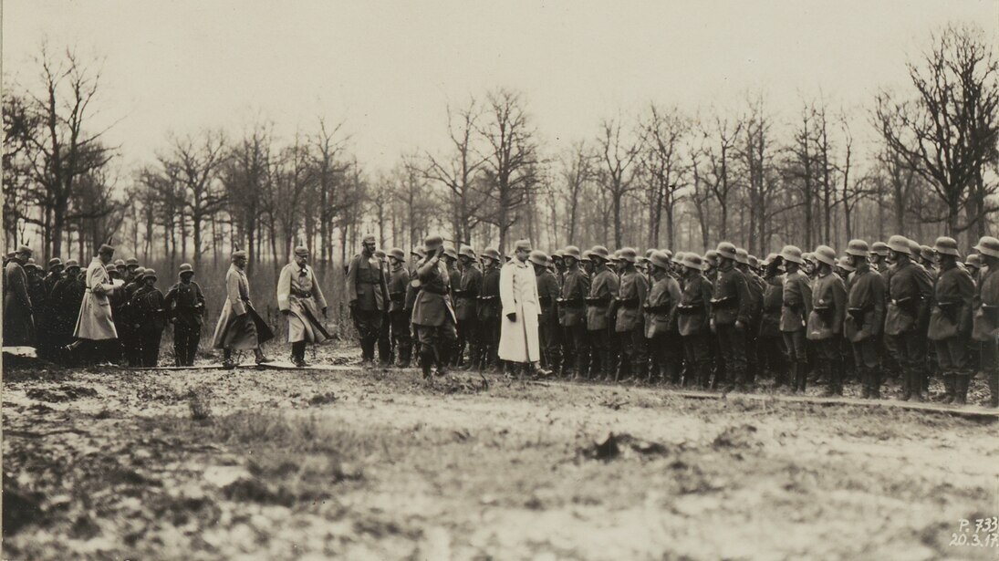 Das Foto zeigt eine Gruppe von Soldaten, vor denen Offiziere entlanglaufen. Im Hintergrund ist eine Reihe kahler Bäume zu sehen.