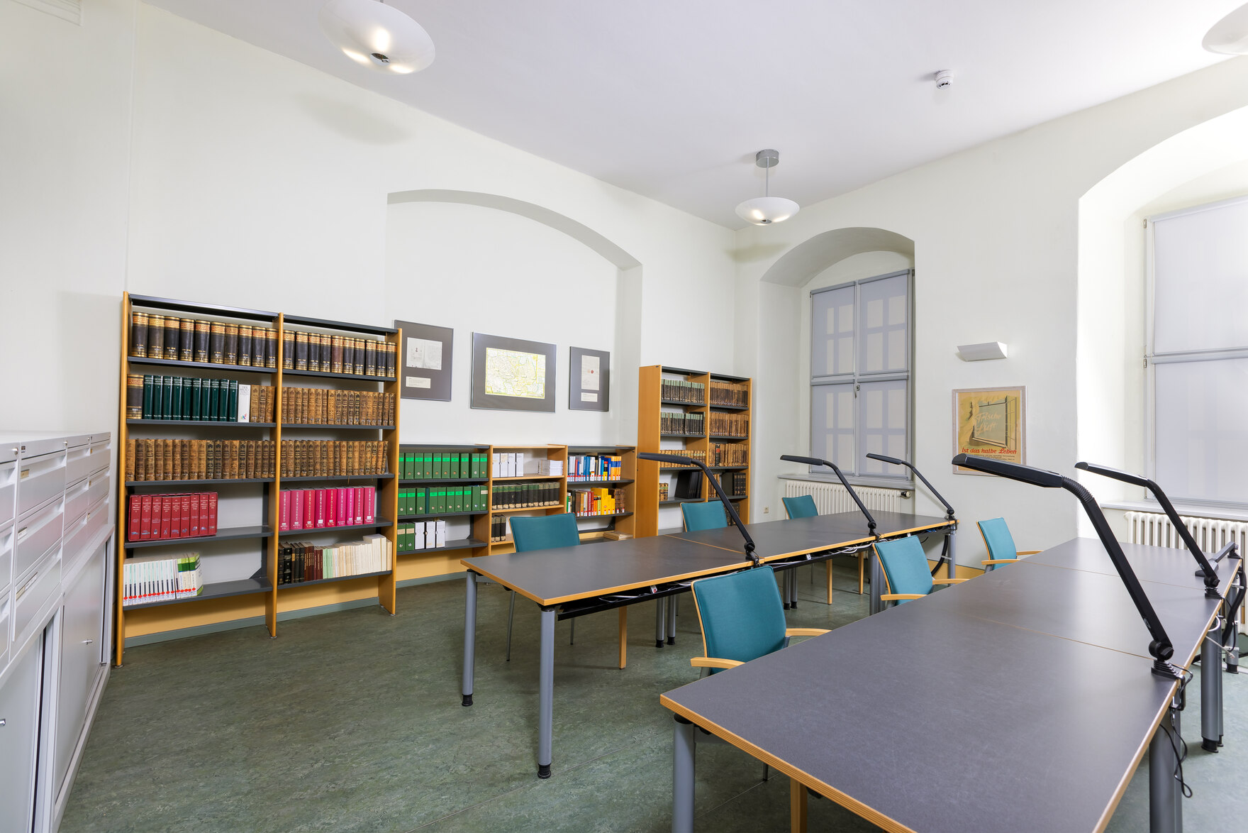 Das Foto zeigt den Lesesaal mit Arbeitstischen im Vordergrund und Bücherregalen an der Wand.