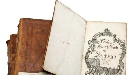 Bücherstapel, das oberste Buch ist aufgeschlagen. Dessen Titelblatt zeigt einen großformatigen, mit schwarzer Tinte und Feder gezeichneten verzierten Rahmen mit dem Text Tauf- und Handelsbuch zu Grießbach 1765.