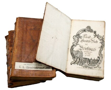 Bücherstapel, das oberste Buch ist aufgeschlagen. Dessen Titelblatt zeigt einen großformatigen, mit schwarzer Tinte und Feder gezeichneten verzierten Rahmen mit dem Text Tauf- und Handelsbuch zu Grießbach 1765.