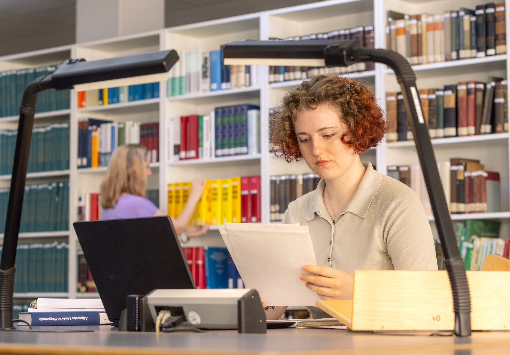 Das Foto zeigt im Vordergrund eine Frau am Arbeitstisch sitzend. Eine andere Frau steht im Hintergrund vor einem Regal.